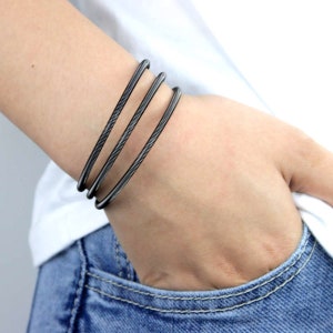 Puede incluir: Un conjunto de tres pulseras de metal negro. Las pulseras están apiladas en la muñeca de una persona.