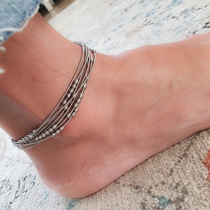 Puede incluir: Un brazalete de tobillo plateado con múltiples hebras y pequeñas cuentas.