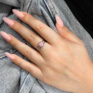 May include: A silver ring with a pink opal gemstone set in a delicate, beaded halo. The ring is worn on a hand with long, light pink nails.