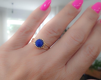 May include: A gold ring with a blue oval gemstone set in a delicate, scalloped bezel. The ring is worn on a finger with pink nail polish.
