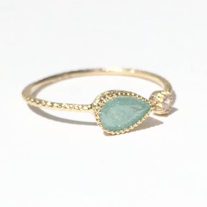 Op de afbeelding: Een gouden ring met een traanvormige groene steen en een kleinere heldere steen. De ring heeft een getextureerde band.