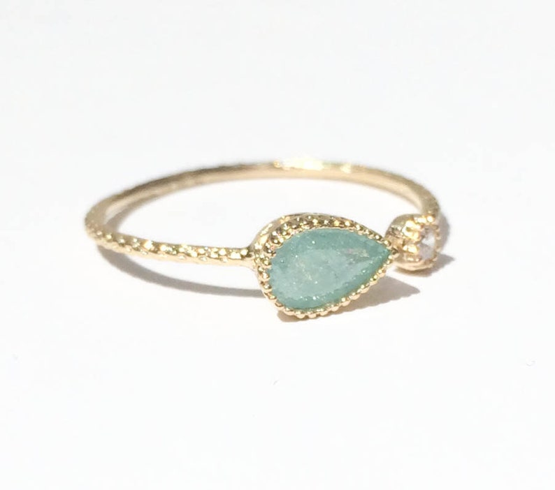 Op de afbeelding: Een gouden ring met een traanvormige groene steen en een kleinere heldere steen. De ring heeft een getextureerde band.