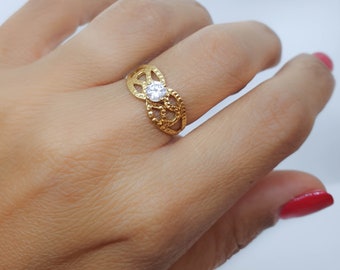 May include: A gold ring with a delicate, intricate design featuring a single, clear, round-cut stone in the center.