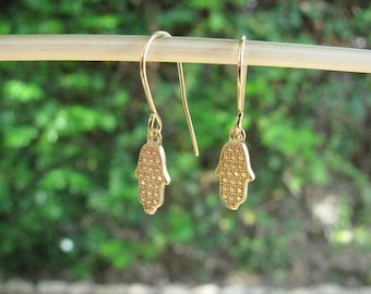 May include: A pair of gold-toned earrings with a hamsa hand design. The earrings are dangle style with a hook closure.