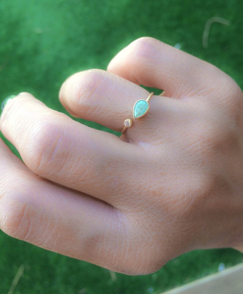 Op de afbeelding: Een gouden ring met een traanvormige groene edelsteen en een kleinere ronde, heldere edelsteen. De ring is open en verstelbaar.
