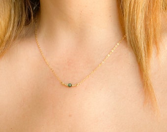 May include: A fine gold necklace with a small, round, green gemstone pendant. The necklace is displayed on a person's neck. The gold chain is delicate, and the gemstone is centrally placed.