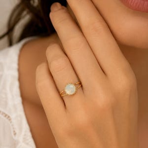 Op de afbeelding: Een gouden ring met een ronde, iriserende witachtige steen die doet denken aan een opaal, gezet in een verfijnde, geschulpte zetting. De ringband is dun en splitst zich in twee parallelle draden bij de aanzet van de centrale steen.