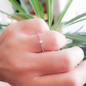 May include: A delicate silver ring with a small, clear gemstone set in the center. The band is adorned with a delicate, intricate design.