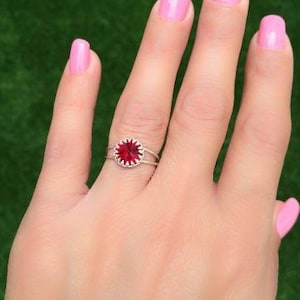 Könnte beinhalten: Ein silberner Ring mit einem roten Edelstein, der in einem kreisförmigen Design gefasst ist. Der Ring wird am Mittelfinger einer Hand mit rosafarbenem Nagellack getragen.
