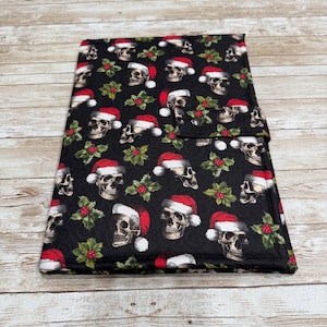 May include: A black fabric item with a repeating pattern of skulls wearing Santa hats and sprigs of holly. The item is rectangular with a flap closure. The background is a light wood grain.
