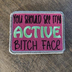 May include: Embroidered patch with the text "YOU SHOULD SEE MY ACTIVE BITCH FACE" in black and green on a pink background. The patch has a silver border and is rectangular in shape.