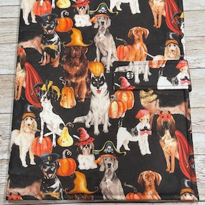 May include: Black fabric notebook cover featuring a repeating pattern of dogs in Halloween costumes. The dogs are wearing witch hats, pirate hats, and capes, with pumpkins scattered throughout the design. The cover has a snap closure.