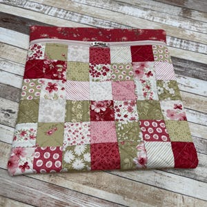 May include: A quilted pouch with a patchwork design. The pouch features a variety of red, pink, and cream floral and patterned fabrics. A red floral fabric lines the top, which has a pink zipper. The pouch is approximately 12 inches wide and 9 inches tall.
