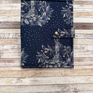 May include: A navy blue journal cover with a white toile pattern featuring the Statue of Liberty and floral elements. The cover has a snap closure with a gold-colored button.