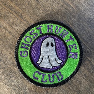 May include: Round embroidered patch with the text "GHOST HUNTER CLUB" in purple on a green background. A white ghost with black eyes is in the center, surrounded by a purple circle. The patch has a black border.