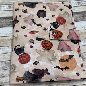 May include: A rectangular fabric cover featuring a beige background with a Halloween-themed print. The pattern includes illustrations of black cats, ghosts, witches on broomsticks, bats, spiders, pumpkins, cauldrons, and candy corn. It has a snap closure.