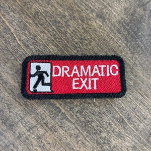 May include: Embroidered patch with a red background and black border. The patch features the words "DRAMATIC EXIT" in white capital letters. A white figure of a person running towards an exit is on the left side of the patch.