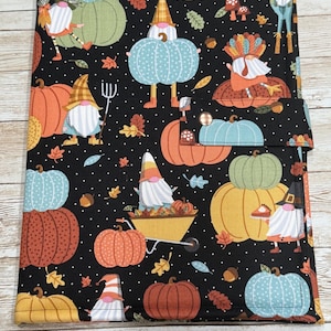 May include: Black fabric cover with a fall-themed pattern. The design features pumpkins in orange, yellow, and teal, along with gnomes, leaves, and acorns. The cover has a flap closure.