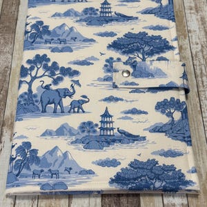 May include: A rectangular fabric cover featuring a blue and cream toile pattern with illustrations of pagodas, elephants, peacocks, and trees. The cover includes a side tab closure secured with a single silver-toned metal snap button fastener.