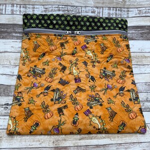 May include: A rectangular quilted pouch featuring an orange fabric with a repeating pattern of pumpkins, owls, and whimsical figures. The top section is a black fabric with a green star pattern, finished with a gray zipper closure at the top.