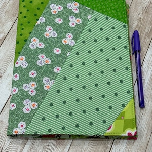 May include: A green fabric binder with a patchwork design featuring different green patterns with white and pink accents. The binder is closed and a blue pen is lying next to it.