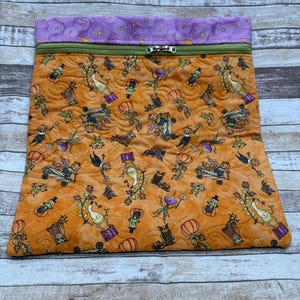 May include: A rectangular fabric pouch featuring a quilted orange exterior with a repeating pattern of whimsical pumpkins, owls, and plant-like figures. The top section is a purple fabric band with a spiral design, separated by a green zipper.