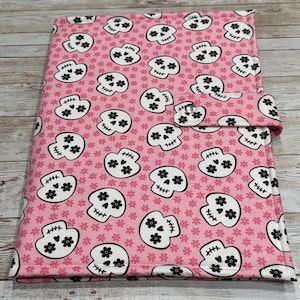 May include: A pink fabric journal cover featuring a repeating pattern of white sugar skulls with black accents and small pink flowers. The cover has a button closure and is designed to protect a notebook or journal.