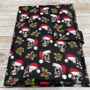 May include: Black fabric item with a repeating pattern of skulls wearing Santa hats and holly sprigs. The item has a flap with a snap closure. The background is a light wood grain.