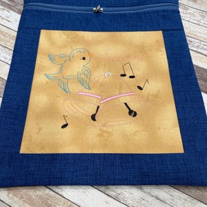 May include: A blue apron with a zipper at the top and a decorative embroidered design. The design features a light blue bird, pink bells, and musical notes on a tan background.