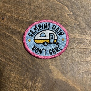 May include: Embroidered patch with the text "Camping Hair Don't Care." The patch features a light blue background with a yellow and white camper design. The border is pink with blue stitching.