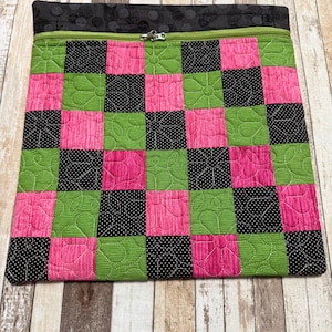 May include: A square quilted pouch featuring a checkerboard pattern of bright pink, lime green, and black fabric squares with white polka dots. The pouch includes a lime green zipper closure and a solid black fabric top border with a subtle texture.