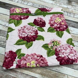 May include: A quilted fabric laptop sleeve featuring a floral hydrangea pattern in shades of pink, burgundy, and yellow, with green leaves. The sleeve has a green zipper and a light pink background.