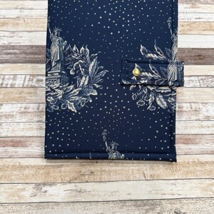 May include: A navy blue fabric journal cover featuring a toile design with the Statue of Liberty, stars, and floral elements. The cover has a flap with a gold button closure. The journal cover is rectangular and appears to be padded.