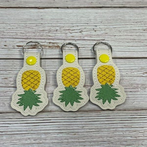 May include: Three white leather keychains with a yellow pineapple design and green leaves. Each keychain has a silver key ring.
