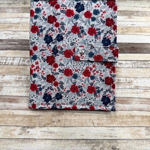 May include: A floral patterned journal cover with a snap closure. The cover features a white background with red and navy blue flowers and green leaves. The journal is closed and resting on a wooden surface.