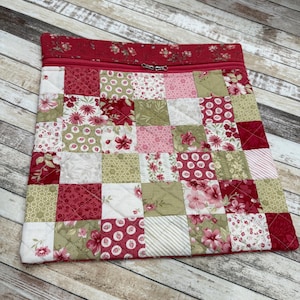 May include: A quilted patchwork zippered pouch featuring squares of floral and patterned fabrics in shades of red, pink, green, and white. The top edge is a solid red with a floral pattern and a zipper closure.