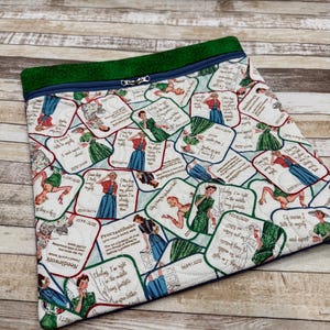 May include: A quilted fabric bag with a green top and a blue zipper. The bag is patterned with vintage-style illustrations of women in dresses, each with a quote. The quotes include "Procrastibake" and "Needlework".
