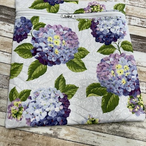 May include: A light gray zippered pouch featuring a repeating pattern of large hydrangea blooms in shades of purple, blue, and yellow, with green leaves. The pouch has a quilted texture and a silver zipper.