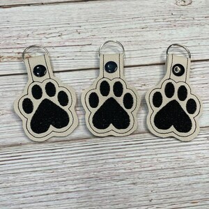 May include: Three cream-colored keychains with black paw print designs. Each keychain has a silver ring.