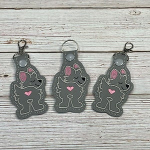 May include: Three gray keychains with a pink embroidered wolf design. Each keychain has a silver metal ring.