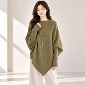 Puede incluir: Un suéter de punto verde oliva con un cuerpo ancho y holgado con dobladillo en punta, mangas largas ajustadas de canalé y un cuello redondo sencillo. Un collar largo y fino de tono dorado con un delicado colgante cuelga al centro.