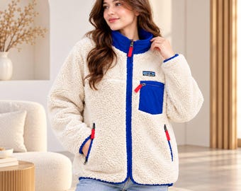 Könnte beinhalten: Eine cremefarbene Fleecejacke mit Hochflor, ausgestattet mit einem leuchtend blauen Stehkragen, blauen Einfassungen und einer Brusttasche. Die Jacke hat einen durchgehenden Reißverschluss mit roten Zippern, einen roten Reißverschluss an der Tasche und ein Patagonia-Logo.