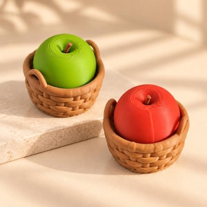 Puede incluir: Dos manzanas pequeñas, redondas y lisas, una de color verde brillante y otra de color rojo intenso, cada una con un tallo marrón fino. Cada manzana está colocada en una cesta circular de color marrón claro con textura trenzada.
