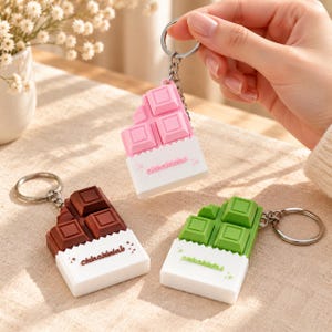 Puede incluir: Tres llaveros con forma de tableta de chocolate en rosa, marrón y verde, cada uno con un envoltorio blanco con el texto chokochok. Los llaveros tienen una anilla y una cadena de metal plateado unidas a la parte superior.