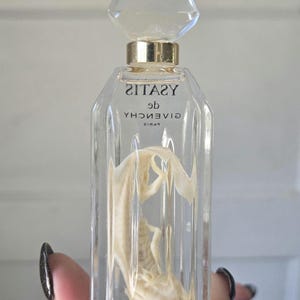 May include: A clear glass perfume bottle with a faceted stopper and gold-tone collar. The bottle features the text YSATIS de GIVENCHY PARIS printed on the front. Inside the bottle, a small, pale, lizard-like specimen is suspended in liquid.