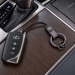 Könnte beinhalten: Ein schwarzer Lexus-Autoschlüssel mit silberner Umrandung und drei Tasten für Verriegeln, Entriegeln und Kofferraum. Der Schlüssel ist über eine schwarze Kordel an einem Metallring mit einem grauen Wildleder-Element befestigt.