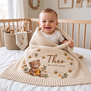 May include: A cream-colored knit baby blanket featuring an embroidered brown bear holding a honey pot, surrounded by small bees, flowers, and the text "Theo Our little honey" in brown script, with a ribbed border edge.