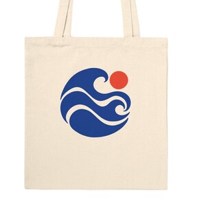 May include: A cream-colored canvas tote bag featuring a centered graphic design of a stylized blue ocean wave and a solid red circle representing the sun. The bag has two matching fabric handles attached to the top edge for carrying.