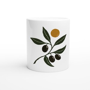 May include: A white ceramic mug featuring a graphic illustration of an olive branch with dark green leaves and three black olives. Above the branch is a solid mustard yellow circle. The design is centered on the smooth white surface.