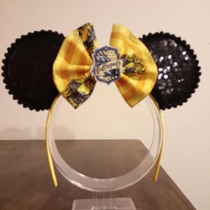 Puede incluir: Diadema con dos orejas circulares de lentejuelas negras y un lazo central de tela a rayas amarillas y doradas. Un pequeño emblema con el texto Hufflepuff está sujeto al centro del lazo. La estructura de la diadema es amarilla y fina.